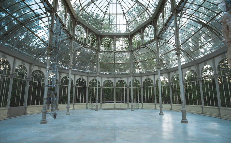Interior view of a large, historic glass conservatory with a domed ceiling made of curved glass panels supported by white metal framework. The floor appears to be made of smooth, light-colored concrete, showing no visible dirt or debris, reflecting the natural light entering through the glass. Tall, narrow windows with arched tops line the perimeter of the room, allowing an abundance of daylight to illuminate the space. The area is clean and well-maintained, with no clutter or dust visible, suitable for surface cleaning and sanitisation. The setting is spacious and bright, ideal for hosting or event planning at Crystal Palace Park, with natural greenery visible through the glass offering a lush backdrop, and no cleaning tools or products are visible in this shot. The overall scene emphasizes the importance of thorough cleaning and maintenance for such elegant, large-scale architectural features, aligning with the services provided by Cleaner Crystal Palace.