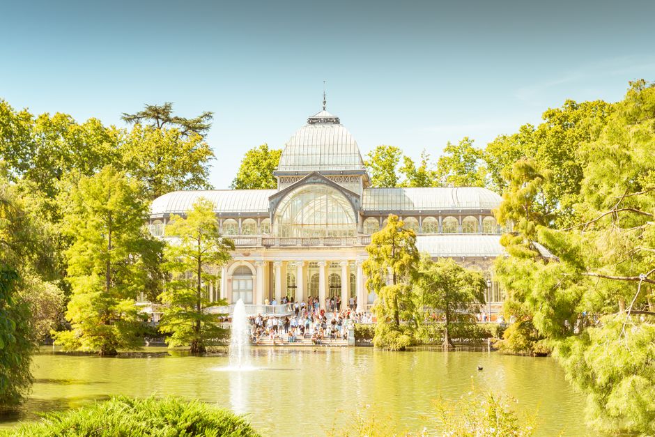 A historic glass and iron conservatory with large, arched windows featuring intricate metal framing and decorative patterns. The glass panels are clear, reflecting the surrounding architecture and clo