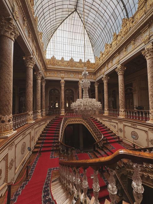 A grand, ornate interior featuring a sweeping red-carpeted staircase with intricate wooden banisters and clear crystal balusters. The room is illuminated by natural light filtering through a large, cu
