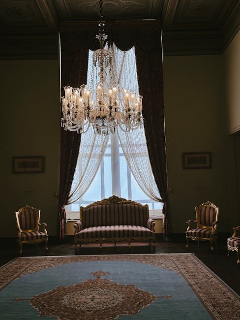A grand, ornate interior featuring a sweeping red-carpeted staircase with intricate wooden banisters and clear crystal balusters. The room is illuminated by natural light filtering through a large, cu